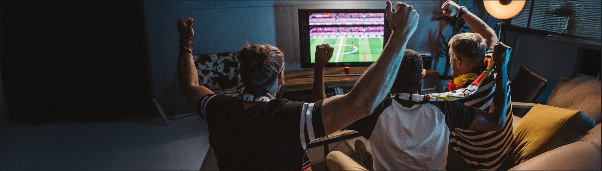 Drei junge Männer sitzen auf dem Sofa und schauen Fußball. Dabei haben Sie Trikots und Fanschals an und jubeln.