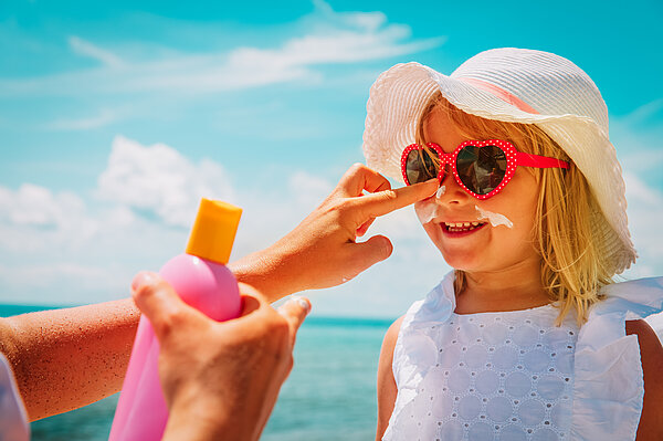Mädchen mit Sonnenbrille und Sonnenhut wird im Gesicht mit Sonnencreme eingecremt. 