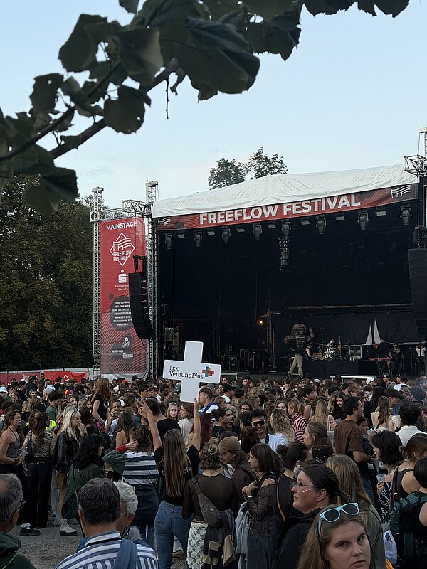 Eine Frau hebt ein großes weißes Plus mit dem BKK VerbundPlus Logo vor der Mainstage des FreeFlow Festivals in die Luft.