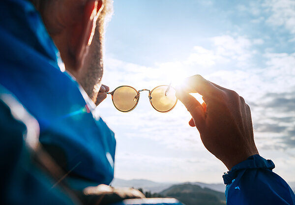 Eine Person hält eine runde Sonnenbrille gegen die helle Sonne und betrachtet den Himmel. Im Hintergrund sind Berge zu sehen, während das Sonnenlicht durch die Gläser scheint.