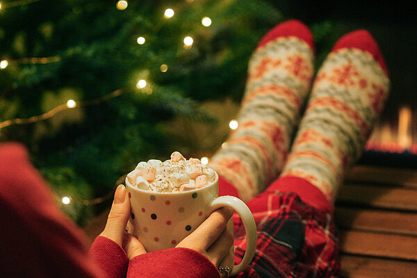 Frau sitzt mit Weihnachtssocken vor dem Christbaum und hält Tasse mit heißer Schokolade und Marshmallows.