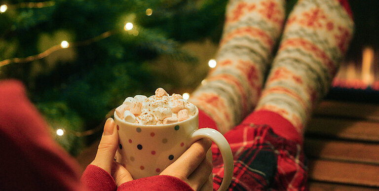 Frau sitzt mit Weihnachtssocken vor dem Christbaum und hält Tasse mit heißer Schokolade und Marshmallows.