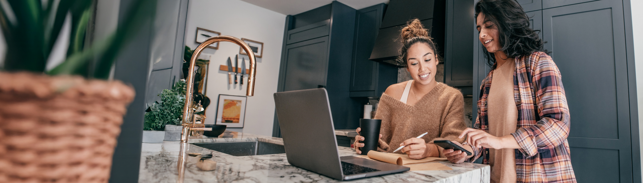 Zwei junge Frauen stehen in der Küche an einem Laptop und machen sich Notitzen.