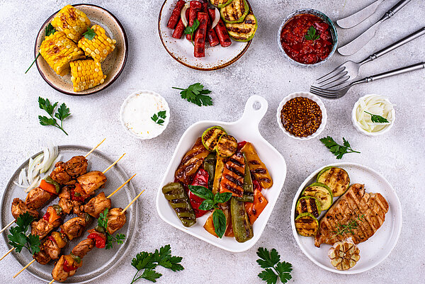 Draufsicht auf verschiedene Grillgerichte: Hähnchenspieße, gegrilltes Gemüse (Paprika, Zucchini), Maiskolben sowie Schälchen mit Dips.