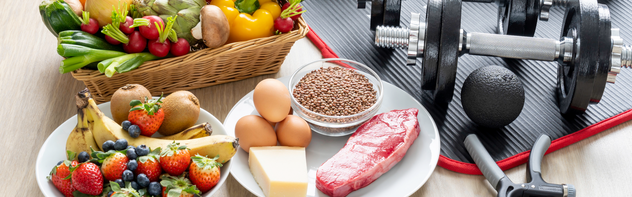 Ein Korb mit Gemüse, eine Schale mit Obst, ein Teller mit verschiedenen eiweißhaltigen Lebensmitteln und Hanteln.