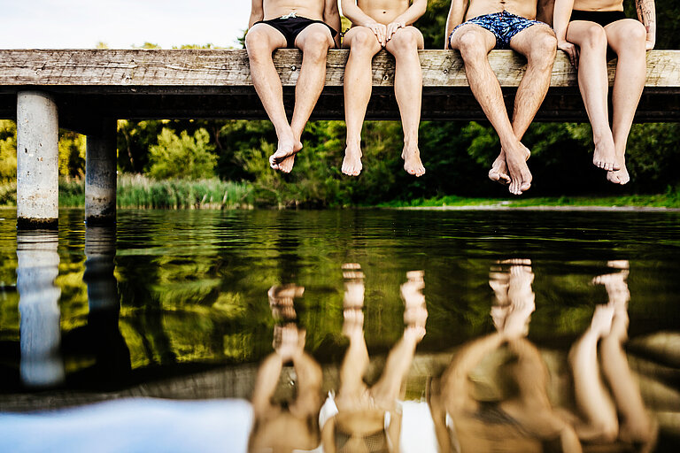 4 Personen sitzen auf einem Steg und lassen die Beine über dem Wasser baumeln. 