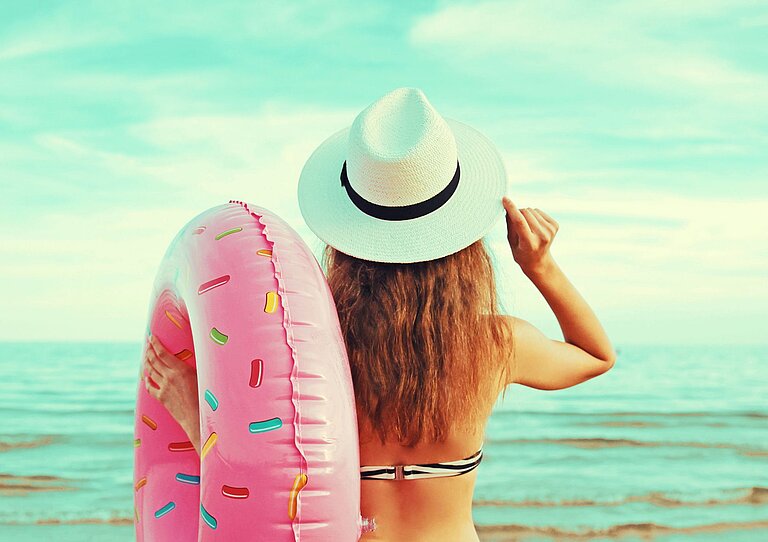 Eine junge Frau schaut auf das offene Meer und hält einen großen Schwimmreifen im Arm.