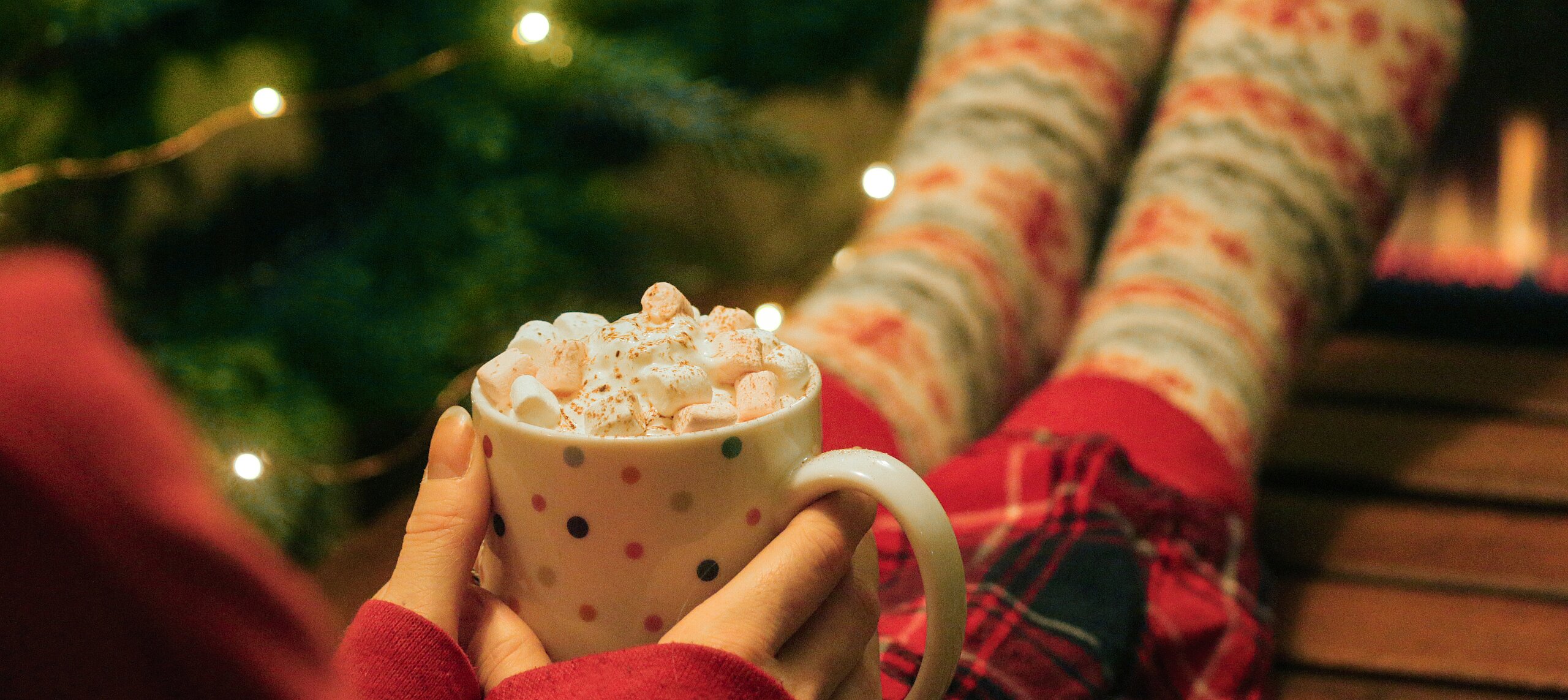 Frau sitzt mit Weihnachtssocken vor dem Christbaum und hält Tasse mit heißer Schokolade und Marshmallows.