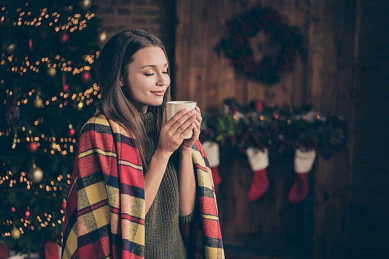 Frau hält eine Tasse in der Hand und hat eine Decke auf den Schultern. Sie steht vor einem Weihnachtsbaum.