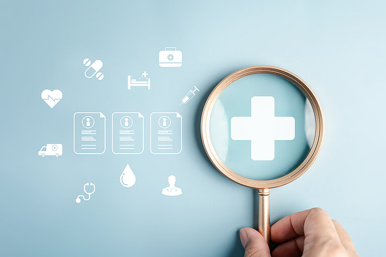 Weiße Krankenkassen- und Gesundheitssystemsymbole auf hellblauem Hintergrund. Durch eine Lupe ist ein Plus-Zeichen zu sehen. 