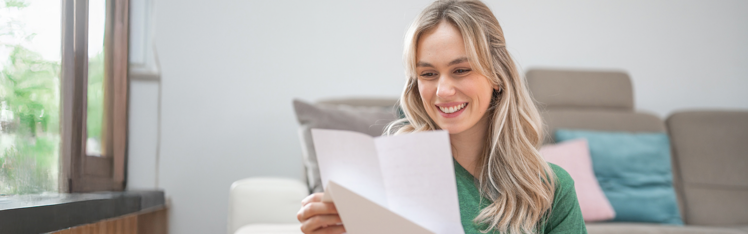 Eine Frau sitzt zu Hause vor dem Sofa und freut sich über einen Brief den sie in der Hand hält