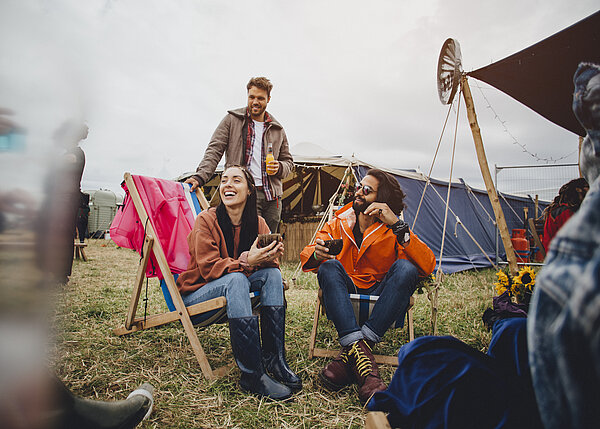Junge Leute beim Zelten, sitzen auf Campingstühlen und lachen. 