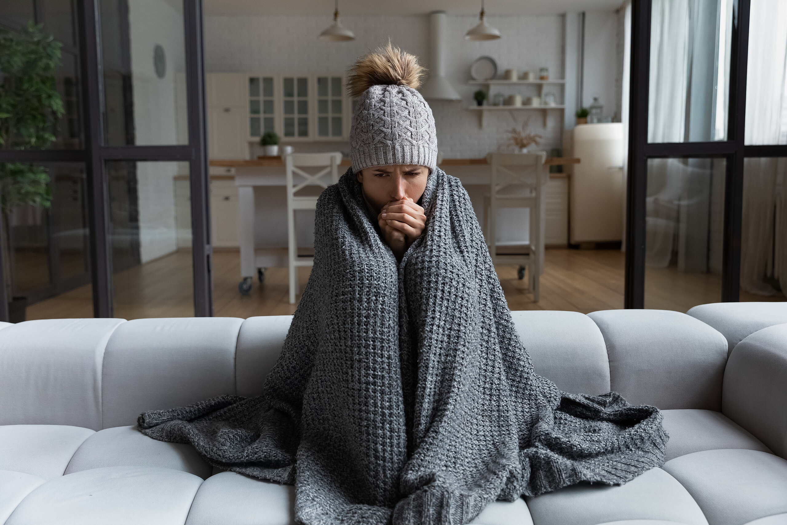 Frau sitzt mit einer Decke um sich und Mütze auf dem Sofa und wärmt sich die Hände.