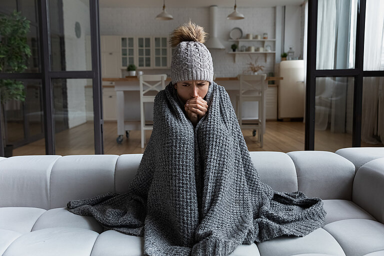 Frau sitzt mit einer Decke um sich und Mütze auf dem Sofa und wärmt sich die Hände.