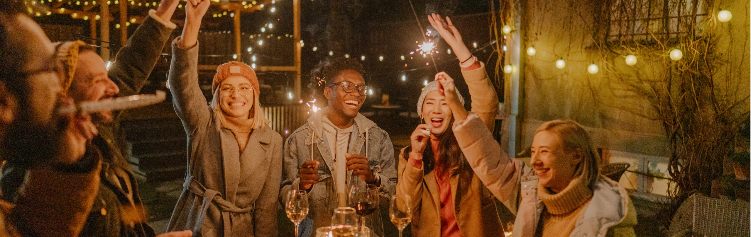 Freundesgruppe feiert Silvester draußen, stehen um einen Stehtisch mit Weingläsern darauf und halten Wunderkerzen nach oben.