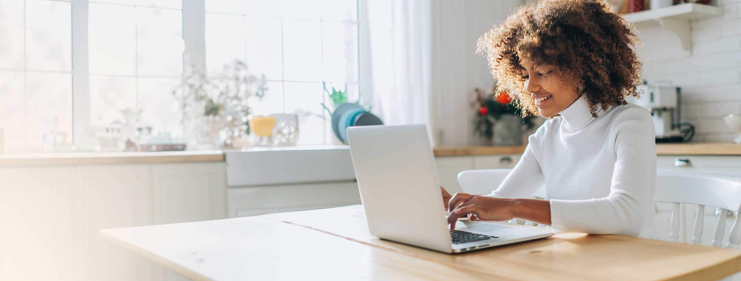 Junge Frau sitzt am Tisch in der Küche vor ihrem Laptop und lächelt.