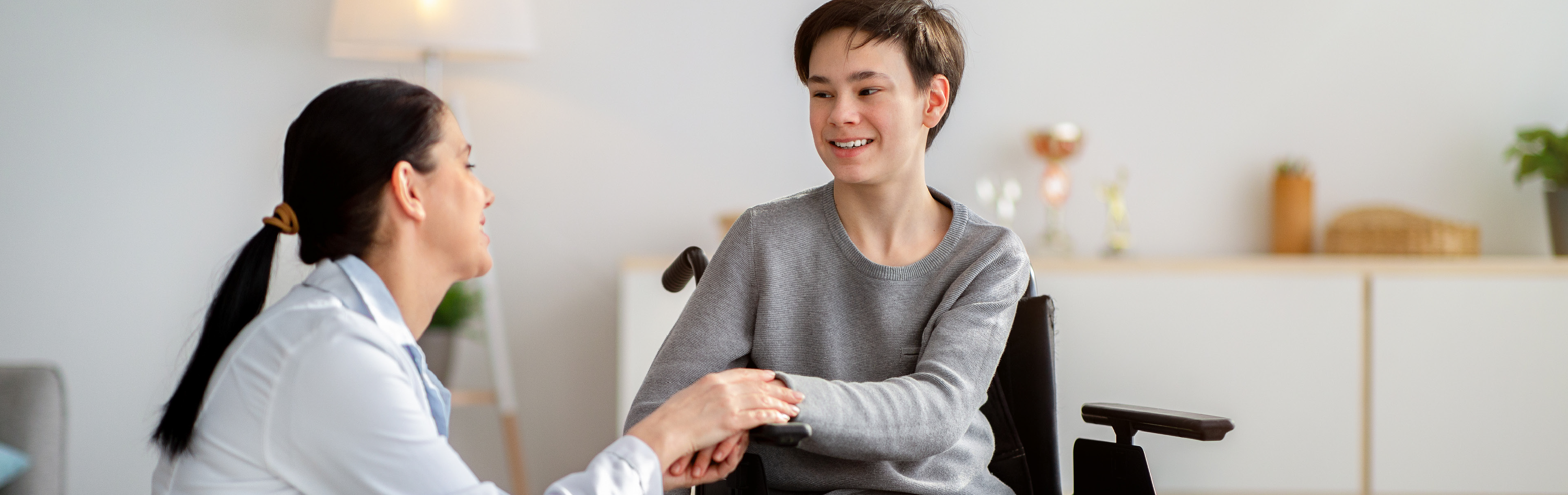 Pflegekraft kniet neben einem Jungen im Rollstuhl und hält die Hand. Beide lächeln.