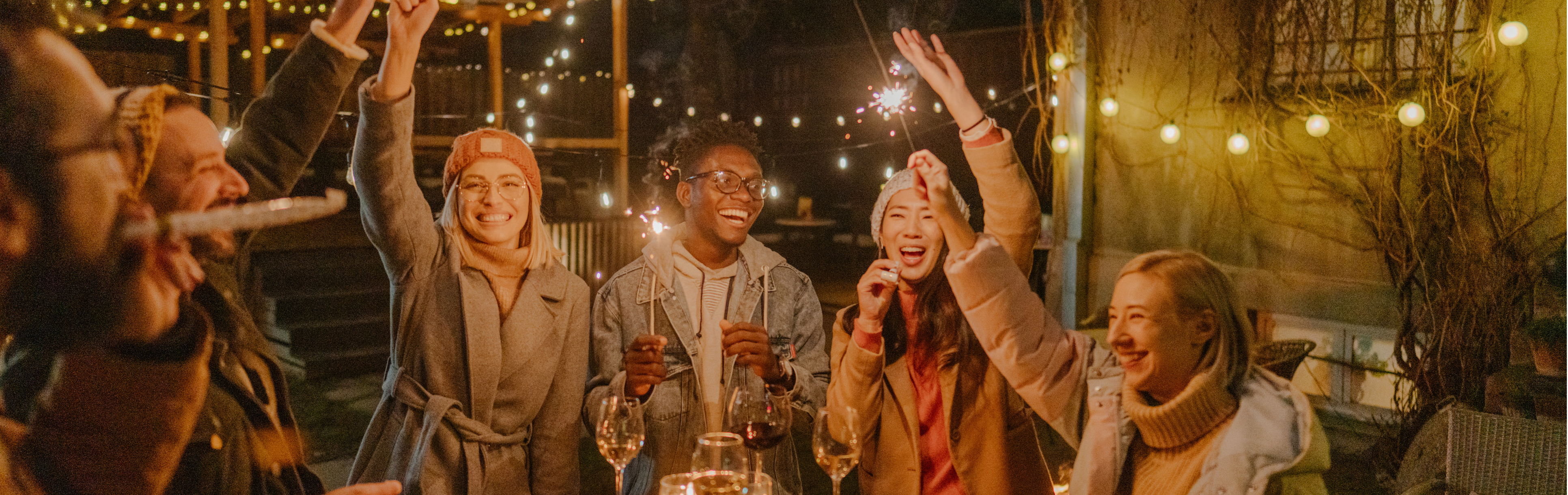 Freundesgruppe feiert Silvester draußen, stehen um einen Stehtisch mit Weingläsern darauf und halten Wunderkerzen nach oben.