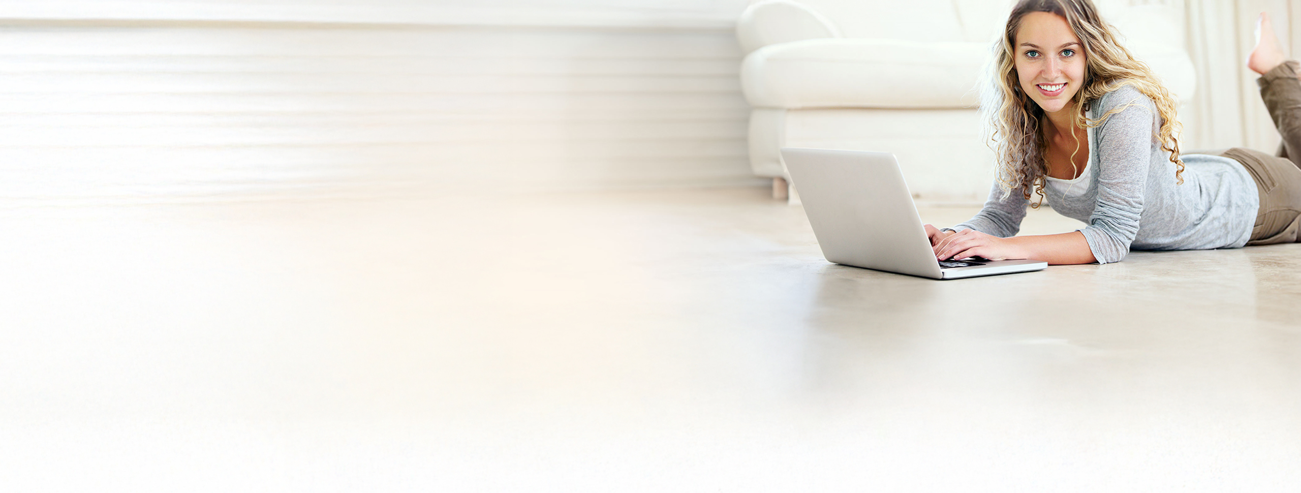 Junge Frau liegt lächelnd mit Laptop auf dem Boden in einem hellen Wohnzimmer.