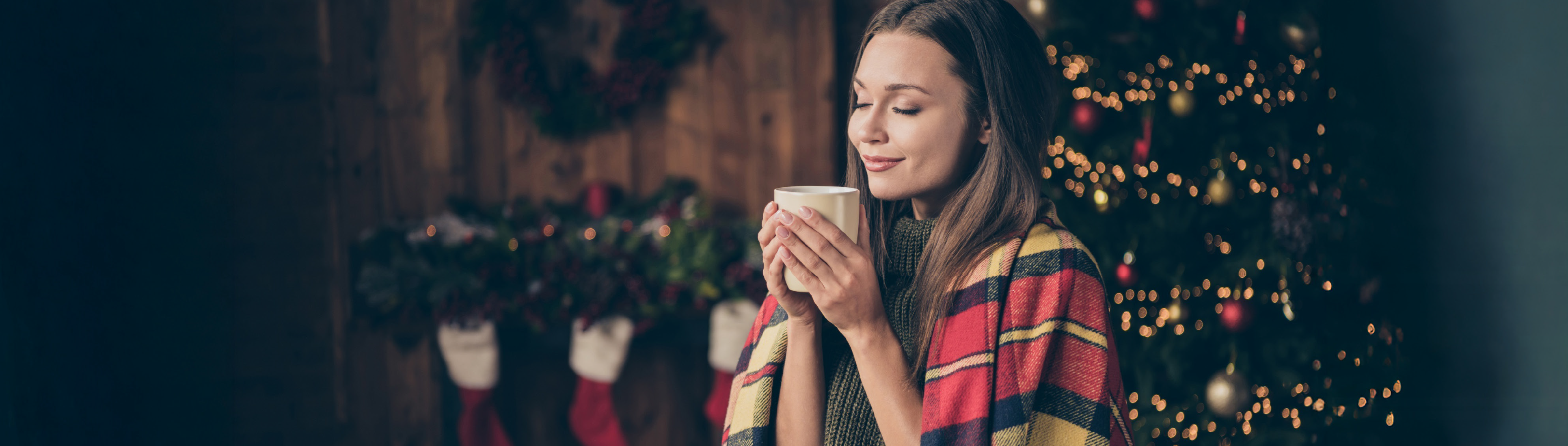 Frau hält eine Tasse in der Hand und hat eine Decke auf den Schultern. Sie steht vor einem Weihnachtsbaum.