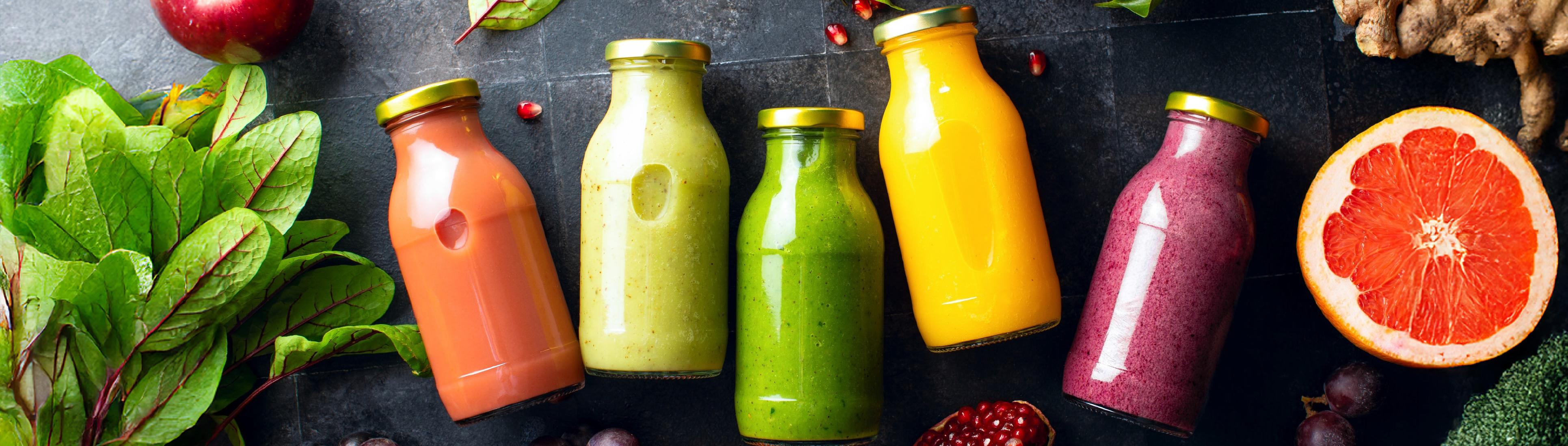 Vier Flaschen, in welcher jeweils ein anderer Smoothie abgefüllt ist. Obst und Gemüse liegen zur Deko drumherum. 