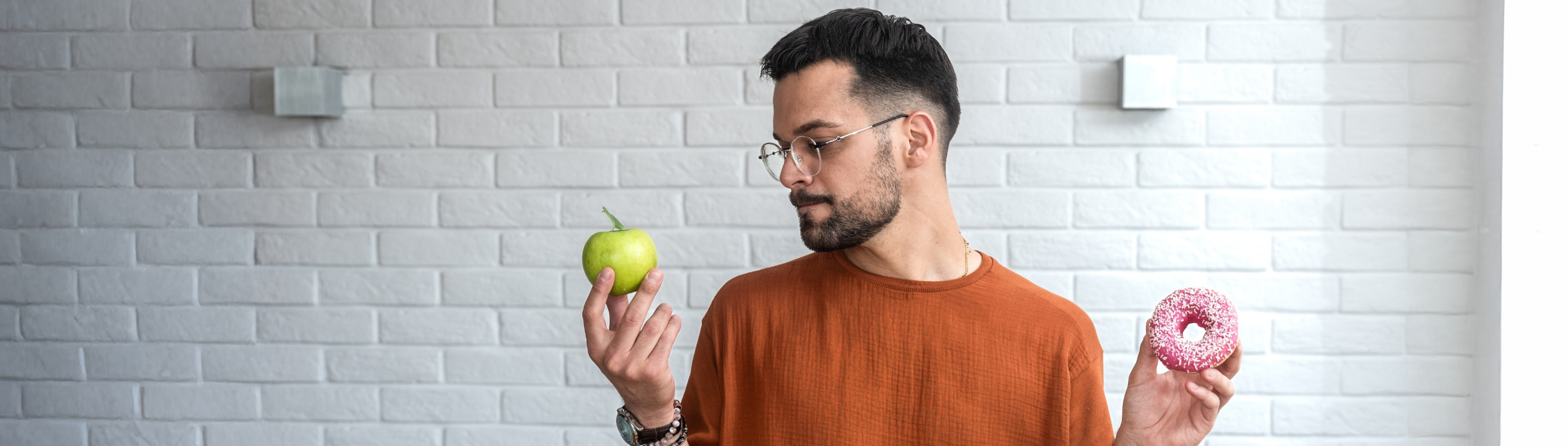 Mann hält einen Donut in der linken Hand und einen Apfel in der rechten Hand. Er schaut den Apfel an.