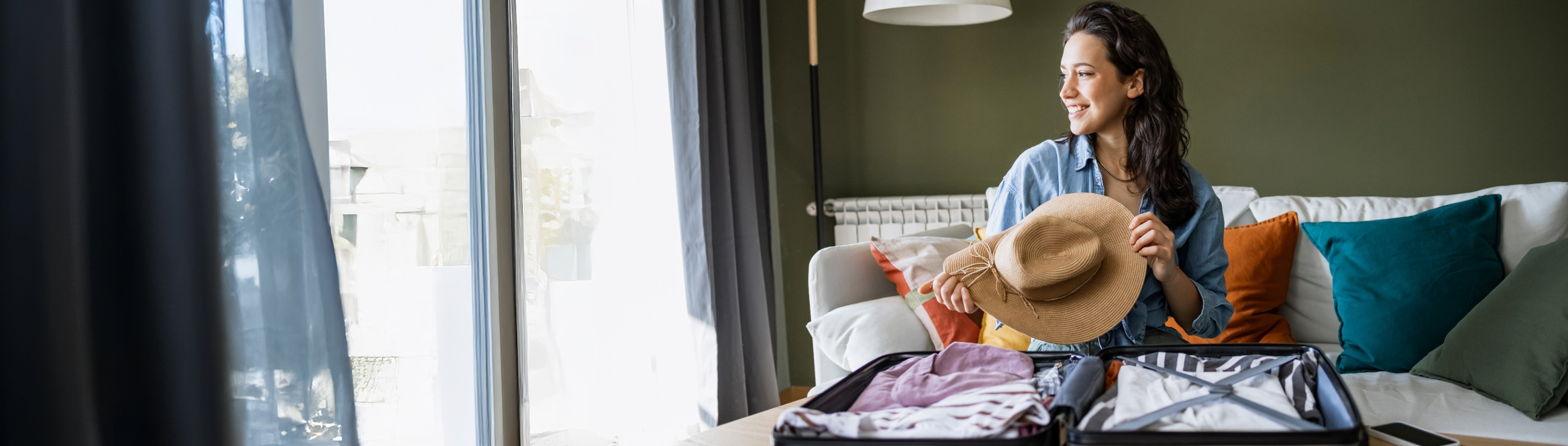 Eine junge Frau sitzt auf dem Sofa, vor ihr liegt ein gepackter Koffer. Sie hält einen Sonnenhut in der Hand und lächelt.