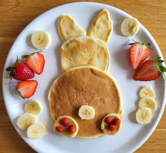 Pancakes, die auf einem Teller liegen und einen Osterhasen darstellen verziert mit Obst. 