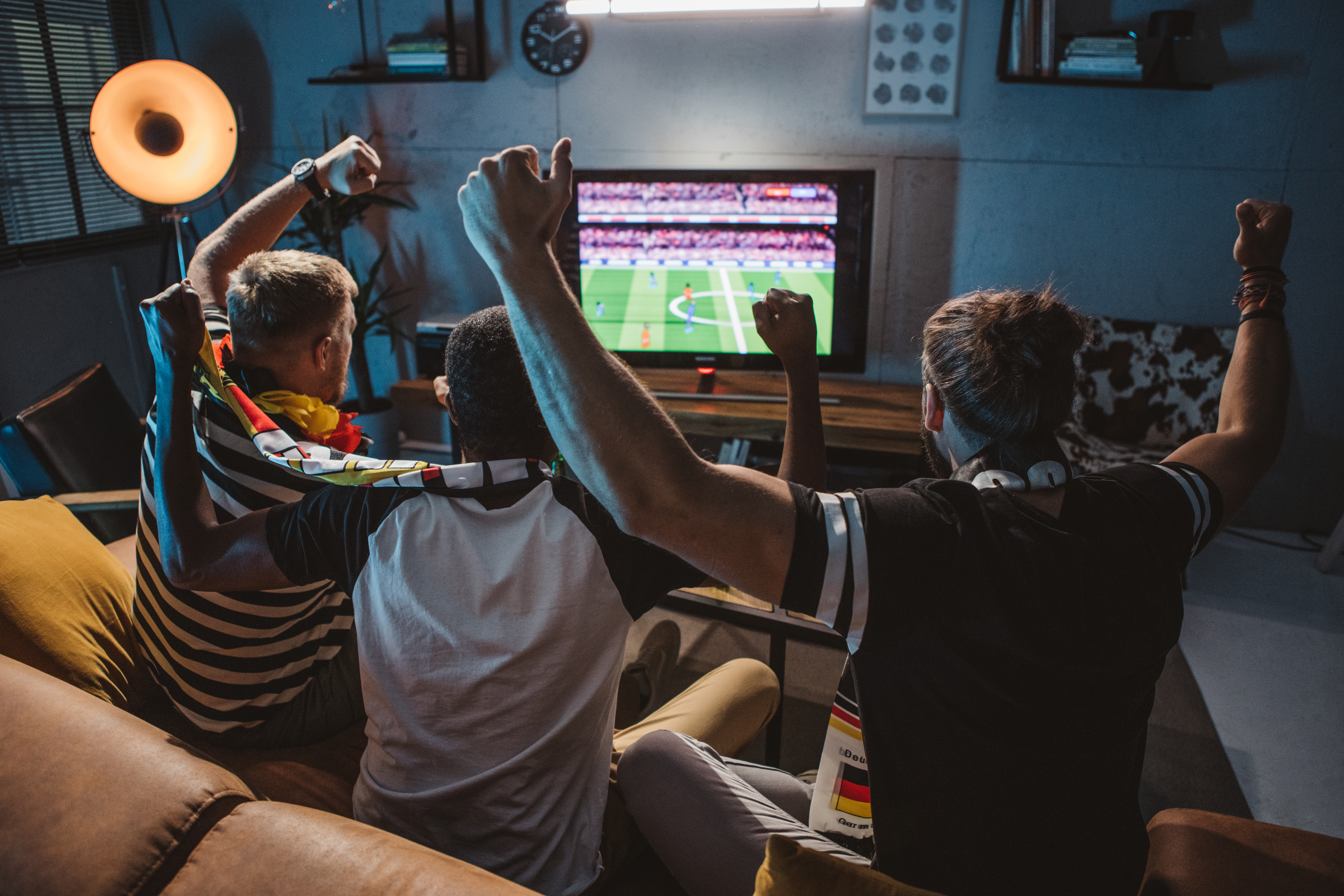 Drei junge Männer sitzen auf dem Sofa und schauen Fußball. Dabei haben Sie Trikots und Fanschals an und jubeln.